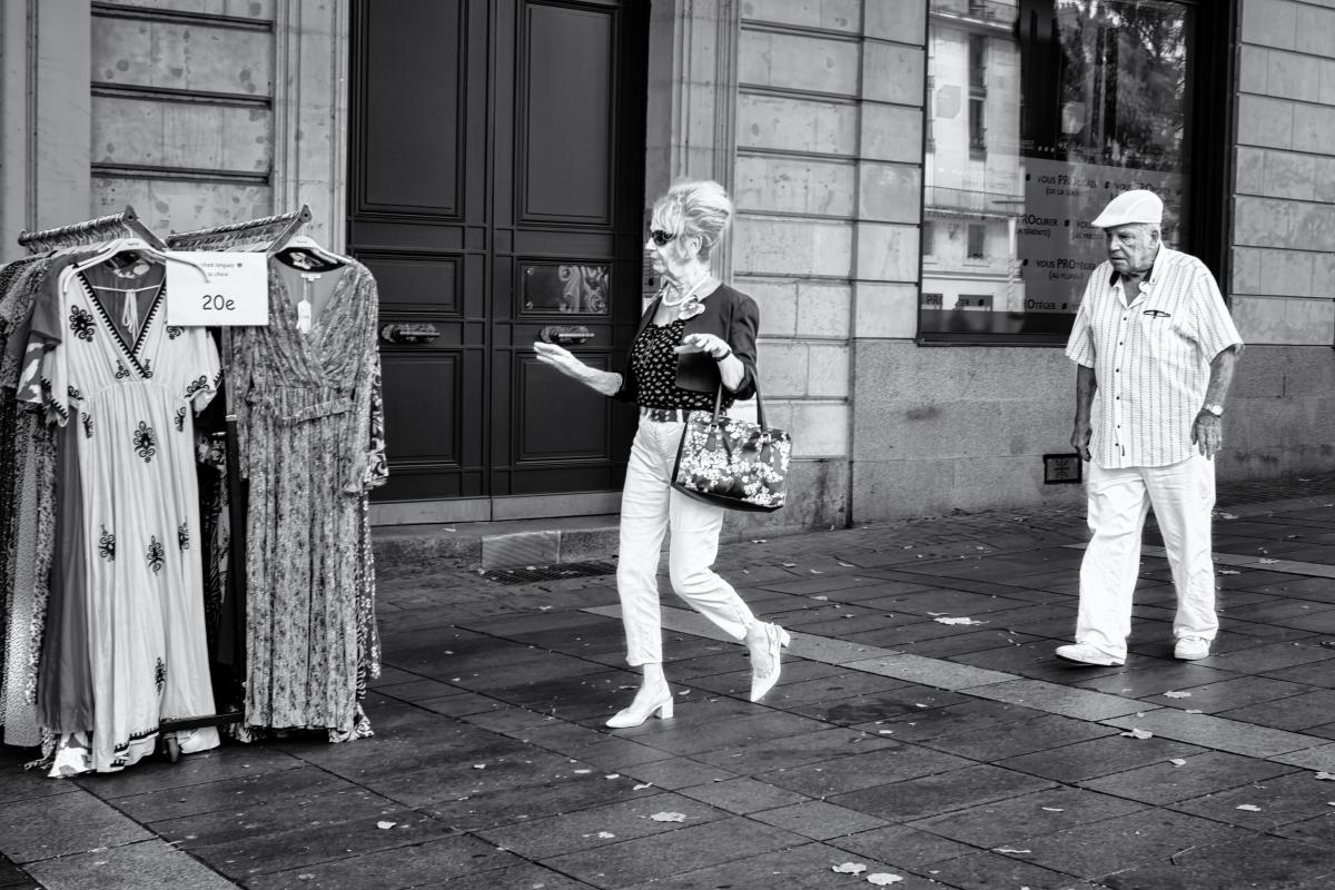 Un homme et une femme âgés et élégants marchent sur le trottoir d'une rue piétonne en pierre, passant devant la devanture d'un magasin et un portant de robes de braderie affichant un prix de 20 euros. La femme au premier plan, portant des lunettes de soleil, un collier de perles et un sac à fleurs, esquisse un geste de la main en regardant vers les vêtements, tandis que l'homme à sa suite, en chemise à rayures et casquette blanche, la regarde marcher. Photographié en noir et blanc très contrasté.